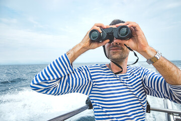 young man, sailor in striped jacket - vest with stubble sits on boat in sea and looks ahead through binoculars. sea navigation, summer tourism. travel content, selective focus