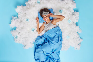 Blissful dark skinned woman lies under blanket on soft pillow smiles gladfully feels rested after healthy enough night sleeping poses on white fluffy cloud against blue background. Above shot