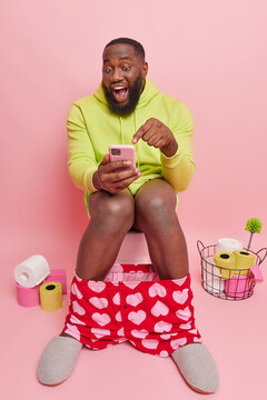 Positive funny bearded man with dark skin points at smartphone display sees something funny defecates on toilet seat dressed in domestic clothes isolated over pink background. Human necessity