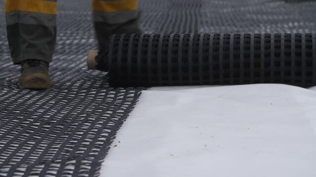 Close-up view of feet of construction worker or builder unrolling roll of plastic net, grid, geogrid or mesh for roadbed reinforcement over road surface. Geosynthetic material for highway base layer.