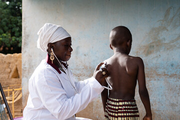Doctor in white uniform placing a stethoscope on the back of a small black boy for measurment of respiratory sounds and heartbeat during a routine health check in West Africa - obrazy, fototapety, plakaty