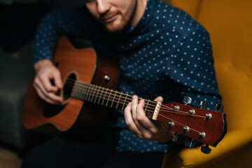 Close-up of guitarist singer male playing acoustic guitar sitting on armchair in dark living room, selective focus. Creative musician enjoying leisure activity in apartment.