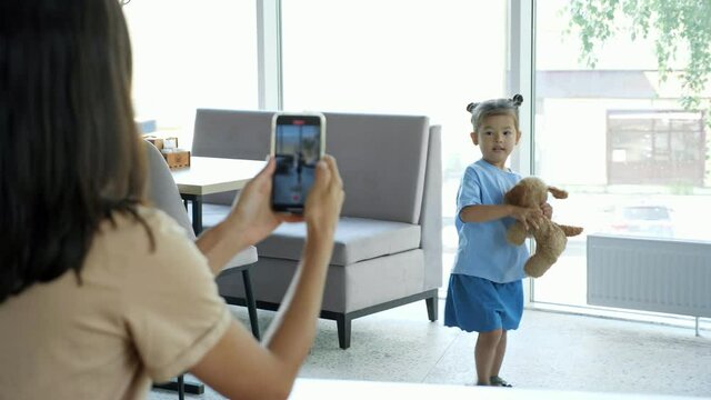 Mom Films Toddler Girl. Woman Brunette Holds Black Smartphone And Makes Photo Of Asian Daughter Playing With Toy Dog And Dancing In Local Cafe Close View