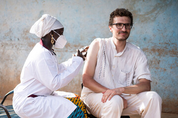 Black nurse wearing a facial mask vaccinating a young caucasian man with a confident smile on his face against yellow fever in an African hospital