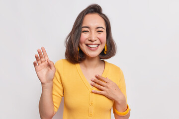 Happy dark haired pretty Asian woman with positive expression laughs joyfully keeps hand raised smiles broadly wears yellow jumper earrings hears something funny isolated over white background.
