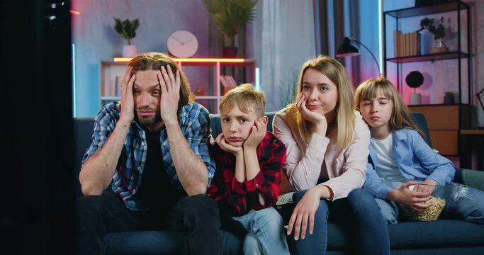 Adorable upset friendly family of four persons-mother,father,son and daughter watching bored film on TV at home in the evening and discussing events between each other