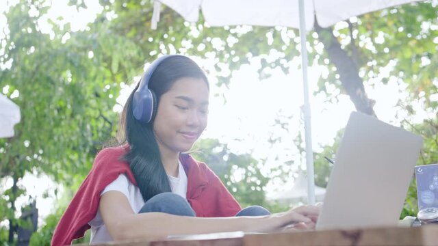 Asian beautiful teenage woman wearing headphones using laptop sits in garden studying online. Video Webinar. remote video conference at home. business online shopping. Work at home. register online