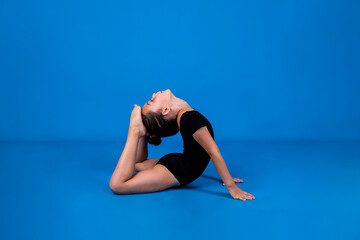 a small gymnast in a black swimsuit does flexibility exercises on a blue background with a copy of the space - obrazy, fototapety, plakaty