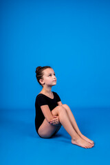 Fototapeta premium a little gymnast in a black swimsuit is sitting on a blue background with a copy of the space