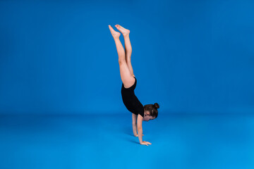 a little gymnast in a black swimsuit stands on her hands on a blue background with a copy of the space
