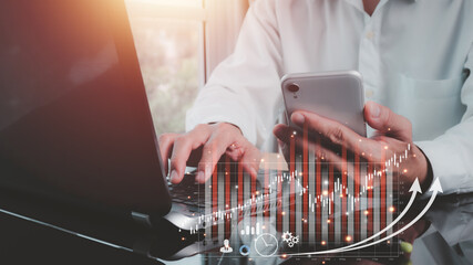 An Asian man in a shirt is sitting at a desk with his left hand holding a mobile phone. His right-hand uses a laptop and a hologram showing a business stock graph. Black background blur