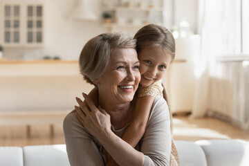 Head shot smiling mature grandmother piggy backing cute little granddaughter, sitting on couch at home, happy middle aged woman with adorable preschool girl having fun hugging, love and gratitude