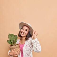 Vertical shot of happy dreamy Asian woman with dark hair buys cactus in pot for her home garden has cheerful expression wears fedora knitted white jumper poses against beige background blank space