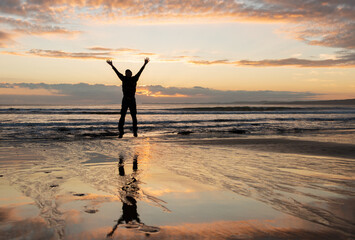 Silhouette man jumping in the air at Milford beach at sunrise with reflection in the wet sand,...
