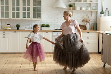 Happy little girl with mature grandmother wearing princess skirts dresses having fun standing in kitchen at home, smiling senior woman with adorable granddaughter enjoying leisure time together