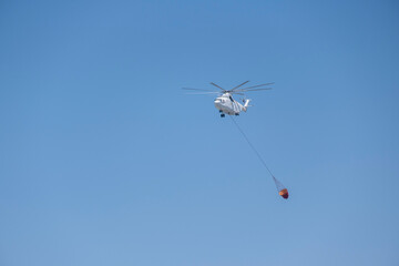 Fire fighting concept: Huge white firefighter helicopter with a long chain carrying red bucket over blue sky on a sunny day. Emergency aircraft background with large copy space.