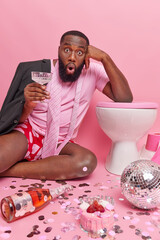 Shocked bearded adult man with thick beard drinks cocktail leans on toilet bowl poses on floor with...