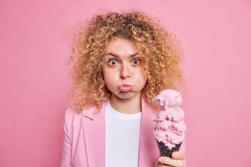 Photo of beautiful curly haired young woman blows cheeks makes funny grimace holds big delicious...