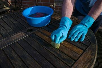 Man with blue rubber gloves is cleaning a dark wooden garden table at the beginning of spring, copy...