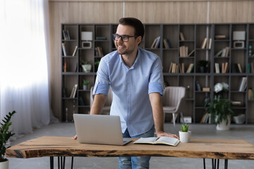 Got an idea. Smiling confident male office worker stand by workplace lean forward to desk with laptop think of achieving success in job. Proud young man student glad to find effective problem solution