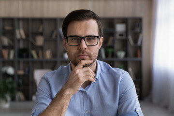 Naklejka premium Thoughtful young man. Headshot portrait of concentrated bearded male teacher analyst look at camera think on idea. Pensive millennial businessman in glasses listen to partner communicate by video call