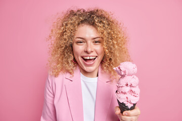 Happy curly haired woman holds big cold ice cream smiles joyfully enjoys summer time and delicious frozen dessert wears formal jacket isolated over pink background. High calorie yummy snack.
