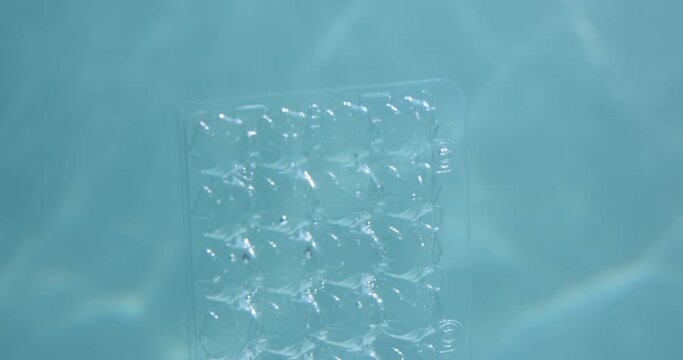 Underwater plastic waste pollution. Close-up discarded egg holder tray floats in light blue ocean background slow motion