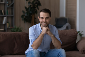 Relax at home. Portrait of happy confident young man student employee sit on sofa at modern living room look at camera enjoy rest on weekend. Positive smiling guy spend free time at comfortable home
