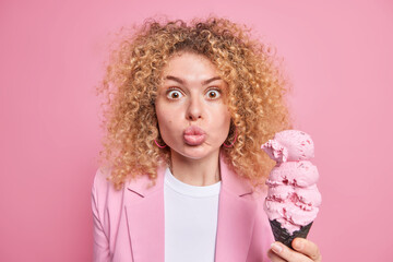 Portrait of surprised woman with curly bushy hair keeps lips folded stares at camera holds big appetizing cone ice cream in waffle wears formal jacket isolated over pink background. Summer dessert