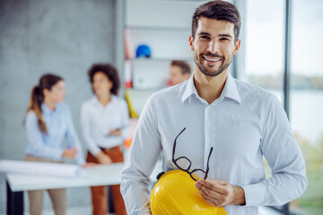 Smiling male architect holding helmet and eyeglasses while looking at camera. There are his colleagues in office working in background.