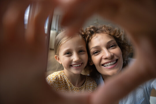 Head Shot Portrait Smiling Mother And Little Daughter Taking Selfie Together, Looking Through Fingers At Camera, Making Heart Shape Gesture, Happy Mom And Adorable Girl Kid Bonding, Expressing Love