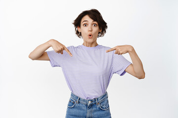 Surprised brunette woman pointing fingers at center, wow face, looking impressed and showing advertisement, sale promo, standing over white background