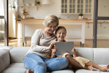 Happy mature grandmother with adorable little granddaughter using tablet relaxing on couch at home together, excited middle aged woman and cute kid looking at device screen, watching funny video