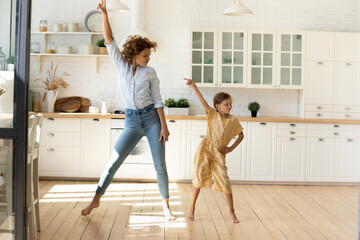 Happy mother and little daughter moving to favorite music in modern kitchen together, young mom...