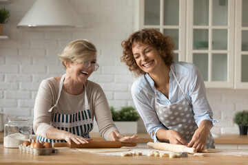 Excited mature mother with grown up daughter having fun, rolling dough together, chatting laughing, cooking homemade cookies or pie, happy woman with senior mom spending leisure time at home