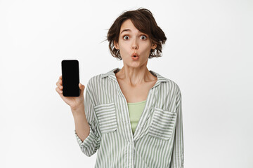 Image of girl looks surprised, shows mobile phone screen, application, shopping app, standing amazed against white background