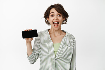 Portrait of surprised brunette woman showing horizontal smartphone screen, looking amazed, standing over white background