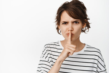 Close up of angry girl hushing, frowning and say shh, press finger to lips, taboo gesture, standing...
