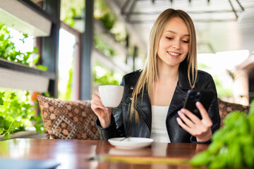 Woman typing write message on smart phone in a modern cafe. CYoung pretty girl sitting at a table with coffee or cappuccino using mobile phone.
