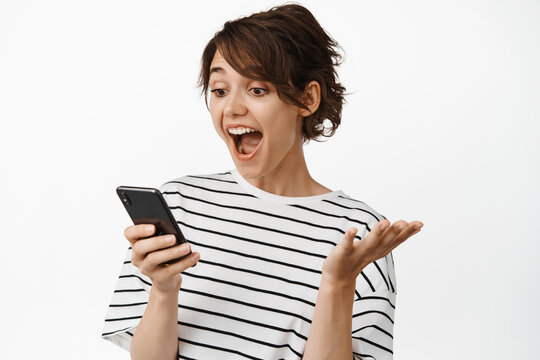 Image of girl looking surprised at mobile phone screen, exciting with great news, notification on smartphone, standing over white background
