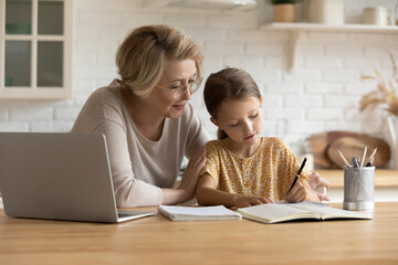 Caring mature grandmother in glasses helping to adorable granddaughter with school homework,...