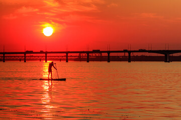 Fototapeta premium Young man on stand up paddle board (paddleboard, SUP) paddleboarding along the Dnieper river at sunset