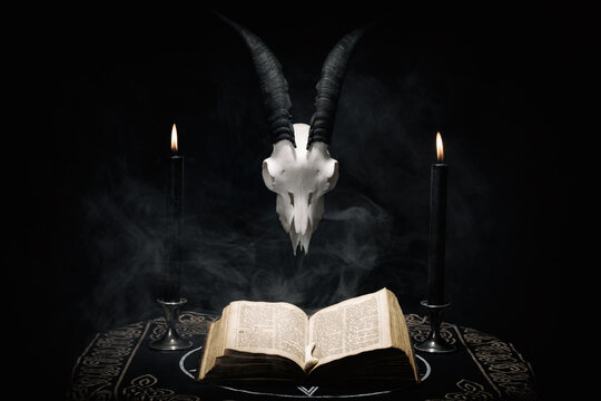 Open old book, goat skull and  black candles on witch table. Occult, esoteric, divination and wicca concept. Halloween background.
