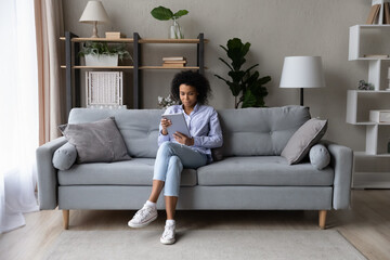 Young African American woman sit relax on couch at home use modern tablet gadget browse internet. Millennial biracial female rest on sofa talk speak on video webcam call on pad device online.