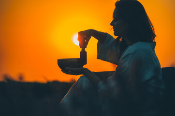 Silhouette of woman playing a Tibetan bowl at gorgeous orange sunset with sun disk framed. Concept:...