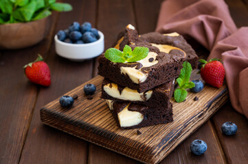 Delicious chocolate brownie cheesecake with fresh berries and mint on a wooden board on a dark wooden background. Copy space.