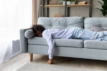 Fototapeta premium Tired young biracial woman lying on couch in living room fall asleep feel fatigue exhaustion. Unwell exhausted millennial African American female relax on sofa sleep or take nap. Tiredness concept.