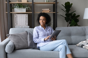 Smiling young African American woman sit relax on sofa in living room use modern tablet gadget look in distance. Happy millennial female rest at home browse internet on pad. Communication concept.