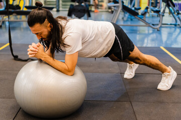 Naklejka premium young hispanic latino man in the gym slim and long-haired doing plank with a swiss ball