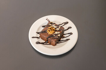 Colorful dessert portion of chocolate brownie cake with dried fruits and nuts, lines of chocolate syrup and white chocolate shavings on a white plate